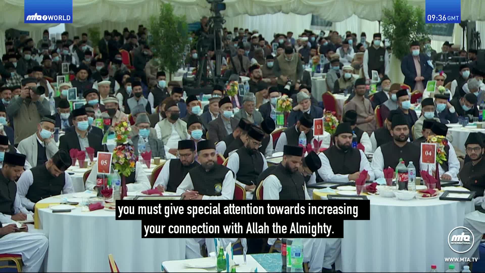 A large crowd sits at round tables, many wearing prayer caps and some with masks. A speaker's words about connecting with Allah are displayed on screen.