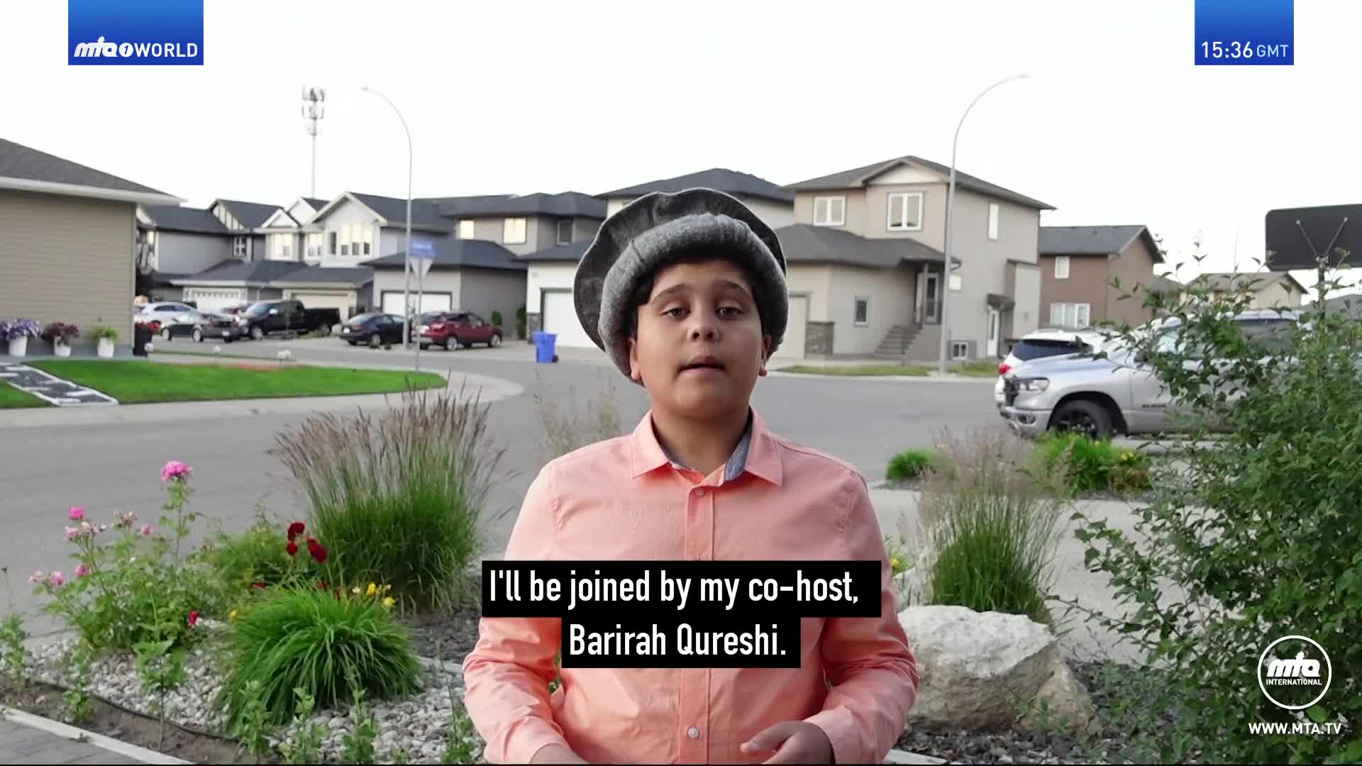 A young boy in a peach shirt and a grey cap stands on a suburban street, speaking directly to the camera. Behind him, houses line a curving road, and cars are parked in driveways.