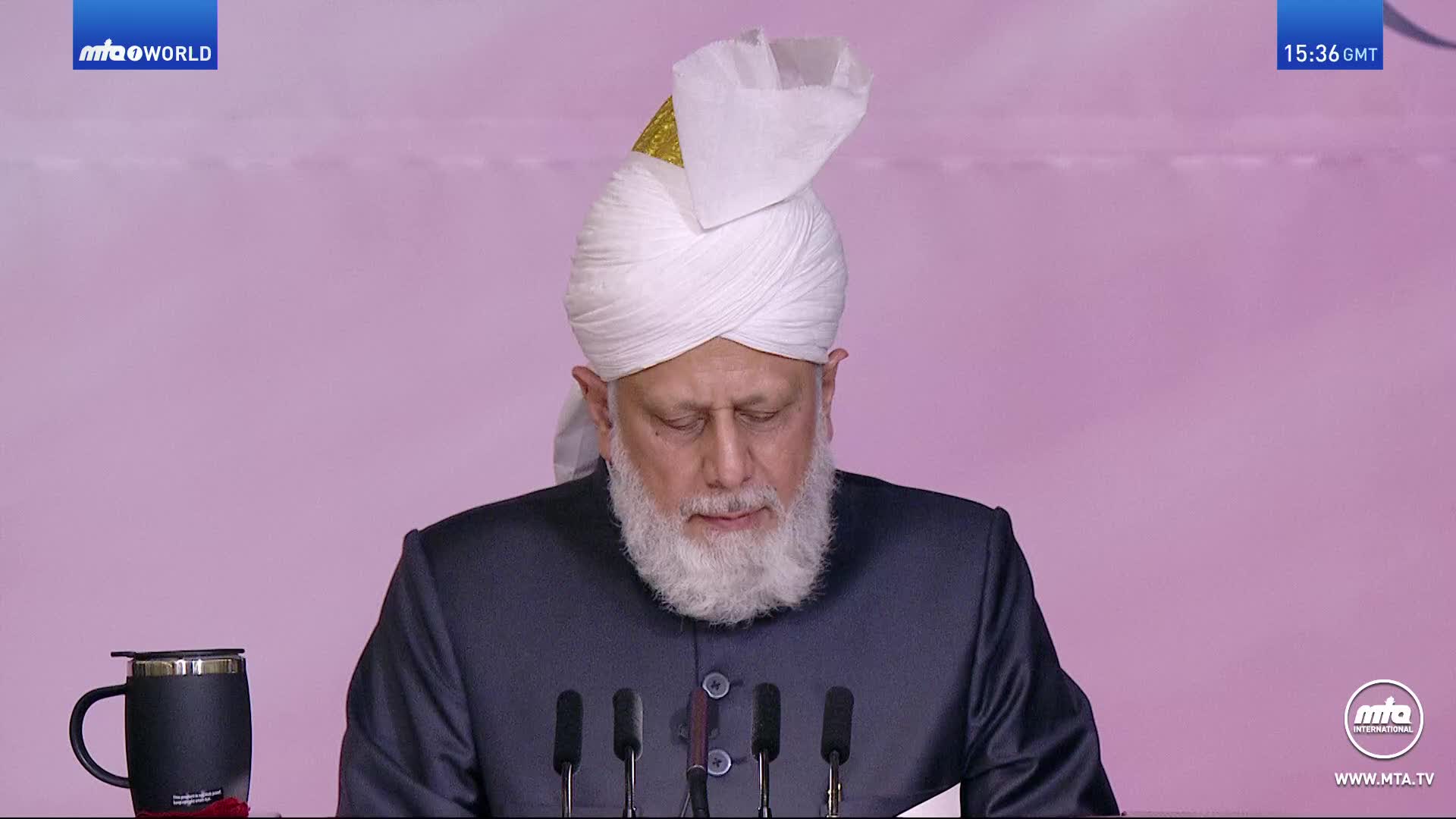 A man in a white turban and dark suit stands before a row of microphones. He looks down, his long white beard prominent. The MTA World logo appears in the top left corner.