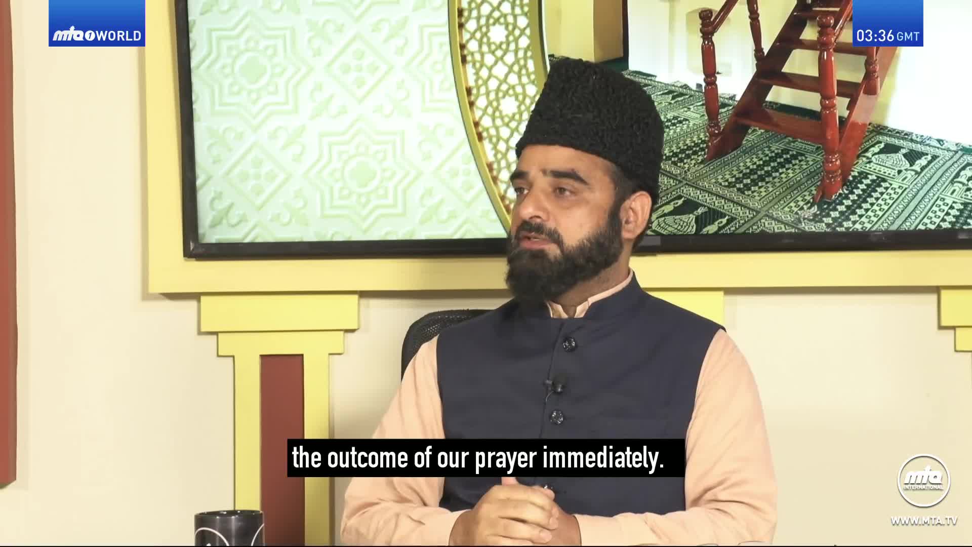 A man in a black cap and vest speaks into a microphone. A screen behind him displays the MTA World logo and a clock showing 03:36 GMT.