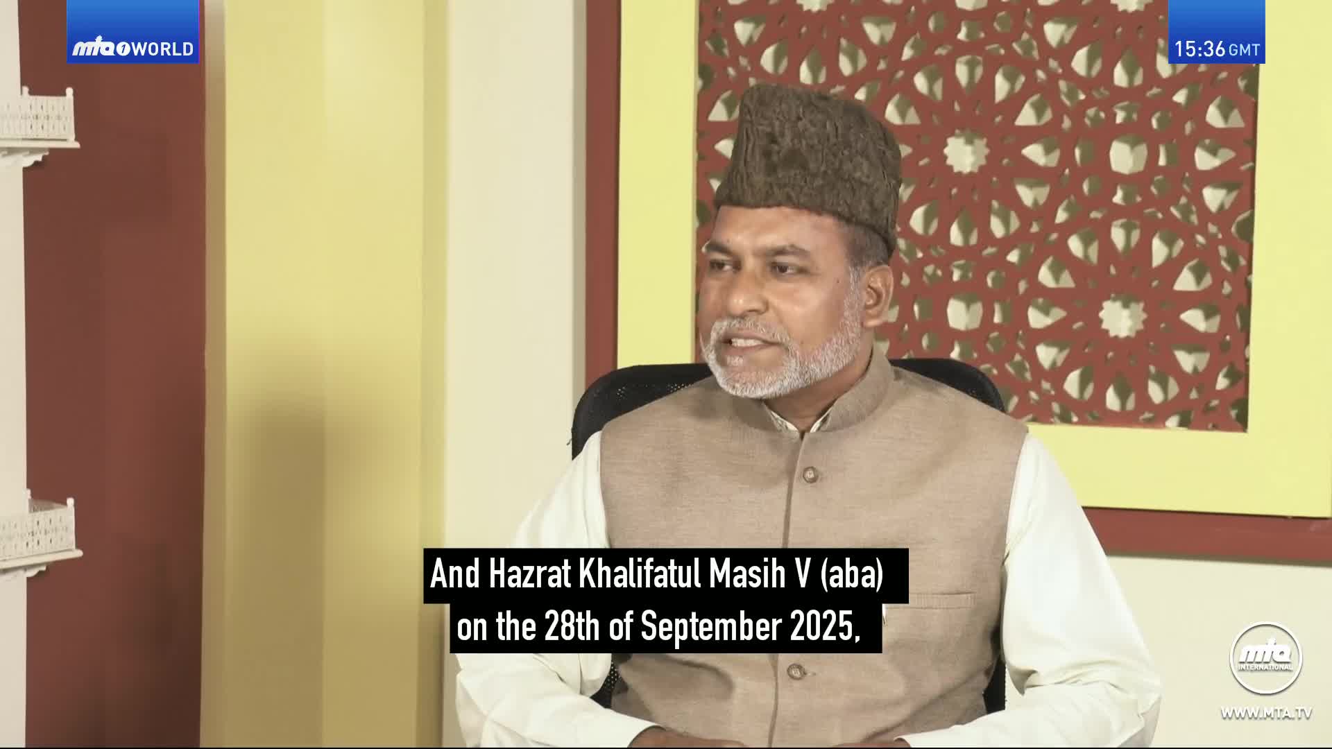 A man with a grey beard and a dark cap speaks, seated in front of a patterned screen. Text at the bottom of the screen reads "And Hazrat Khalifatul Masih V (aba) on the 28th of September 2025."