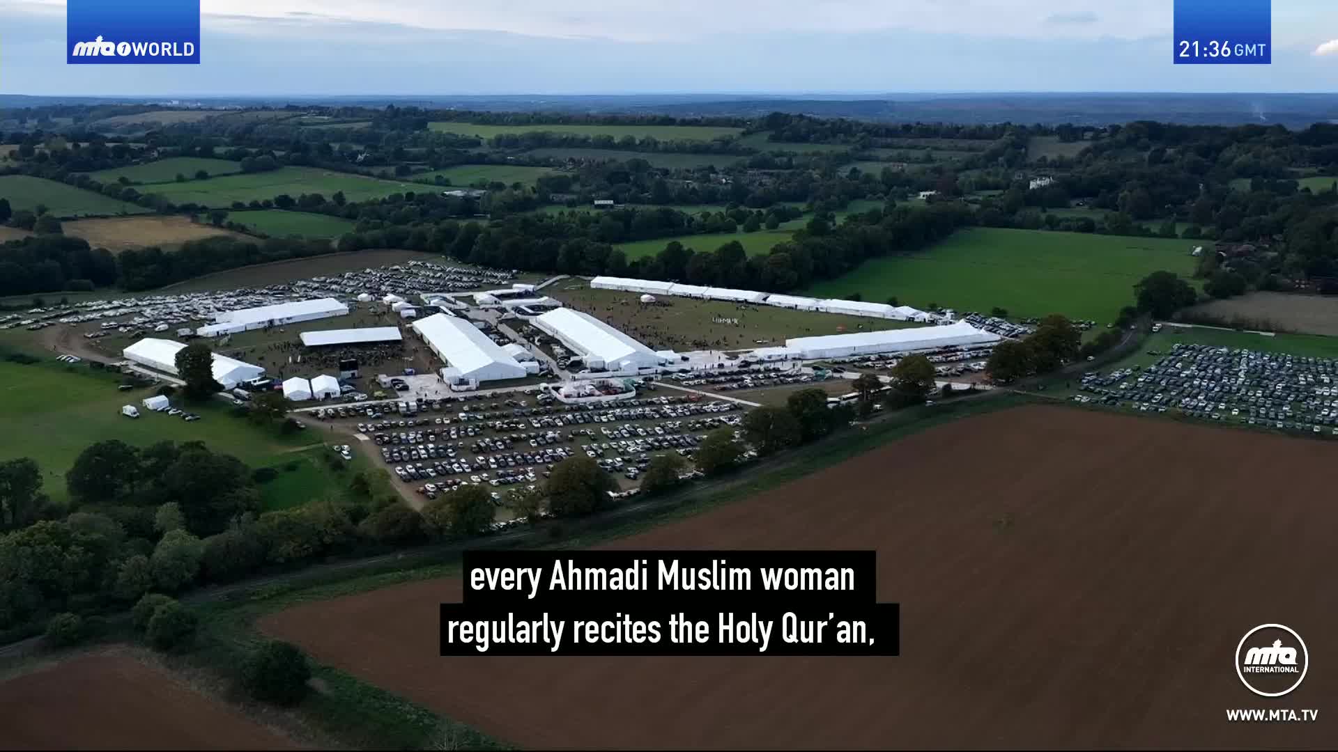 A vast temporary settlement of white tents sprawls across the green fields of the UK countryside. Thousands of cars fill multiple parking areas surrounding the encampment, hinting at a large gathering.