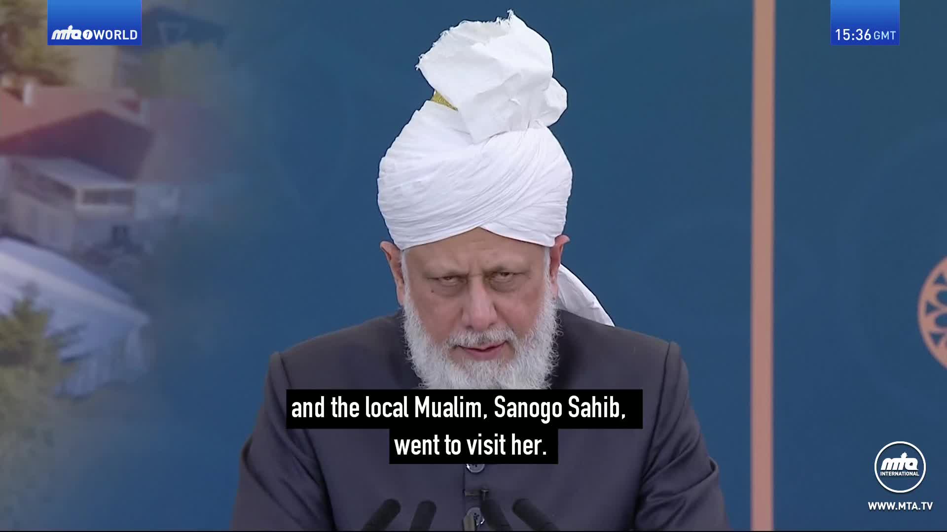 A man with a white turban and beard speaks into a microphone. Behind him, a building is visible on the left, and a blue backdrop with a circular emblem on the right.