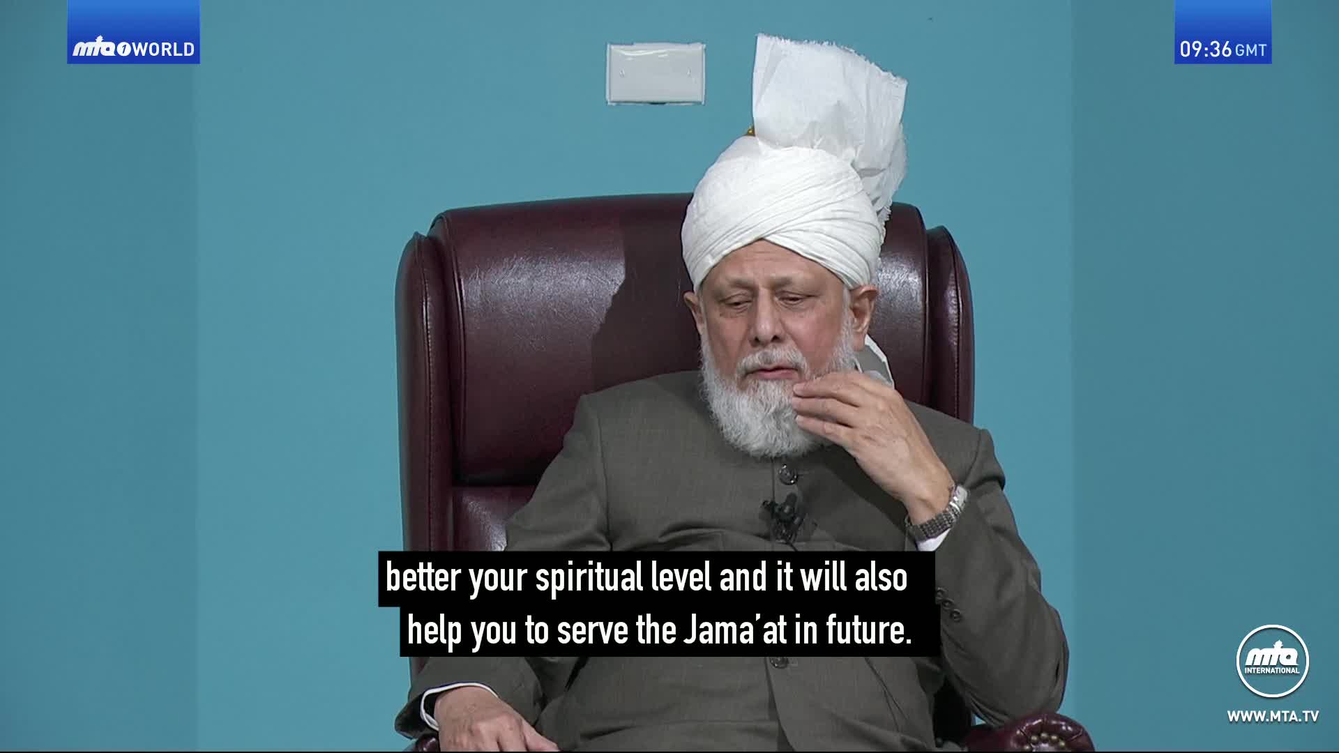 A man in a white turban and grey suit sits in a leather chair, his hand resting on his chin. The screen displays text in English, indicating this is a broadcast from MTA World.