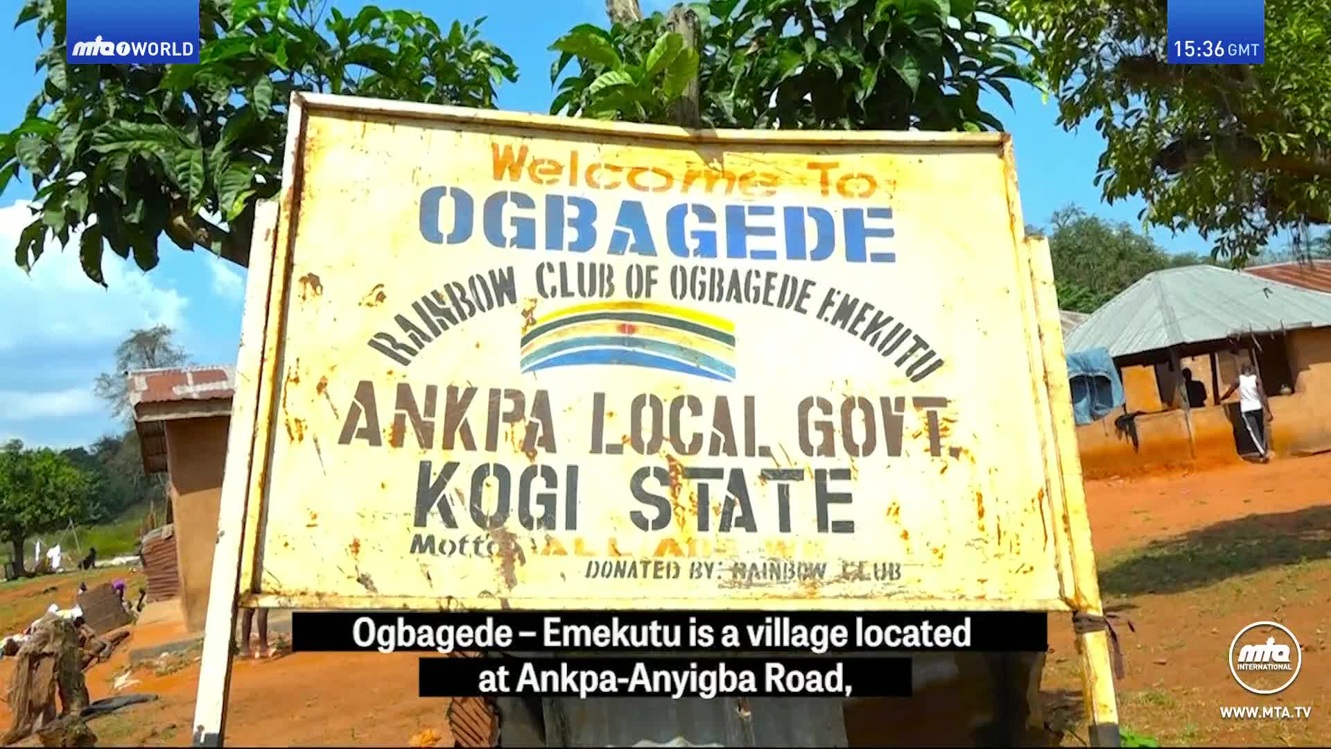 A weathered sign welcomes visitors to Ogbagede, Emekutu, a village in Kogi State. Behind it, a few mud-brick buildings stand under a bright sky.