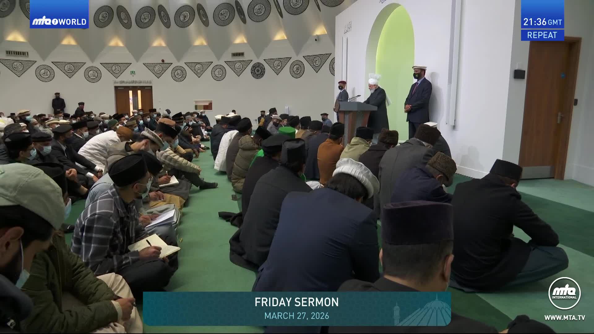 A man in a dark coat stands at a podium, addressing a congregation seated on the floor. Many in the audience are taking notes as the Friday sermon continues.