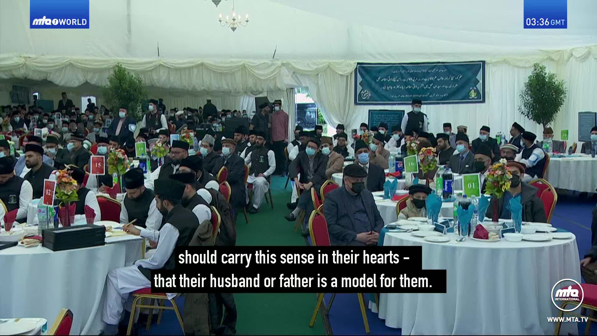 A large crowd of men, many wearing traditional Islamic attire and face masks, are seated at round tables under a white tent. A banner with Arabic script hangs behind them, and the MTA World logo is visible in the corner.