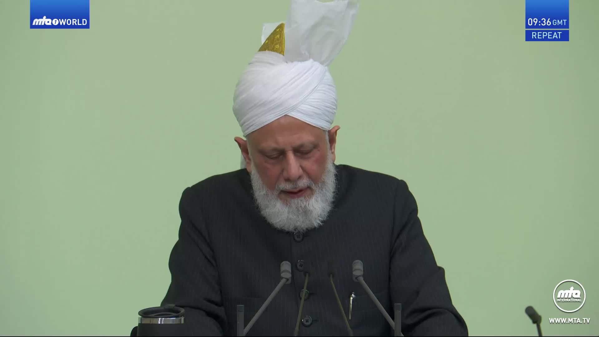 A man in a white turban and dark jacket speaks at a podium. The MTA World logo appears in the top left corner.