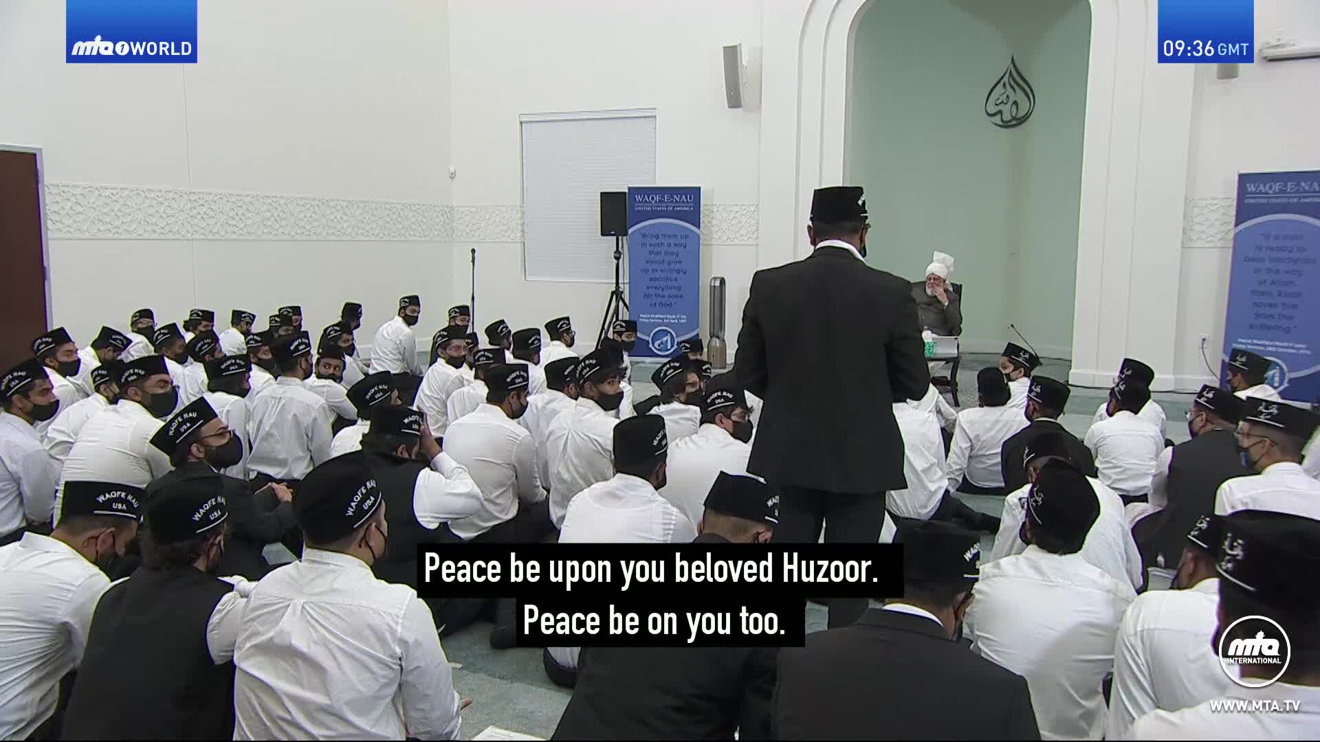 A group of men in white shirts and black hats sit on the floor, facing a speaker. The speaker, also in a black suit and hat, stands and addresses the seated assembly.