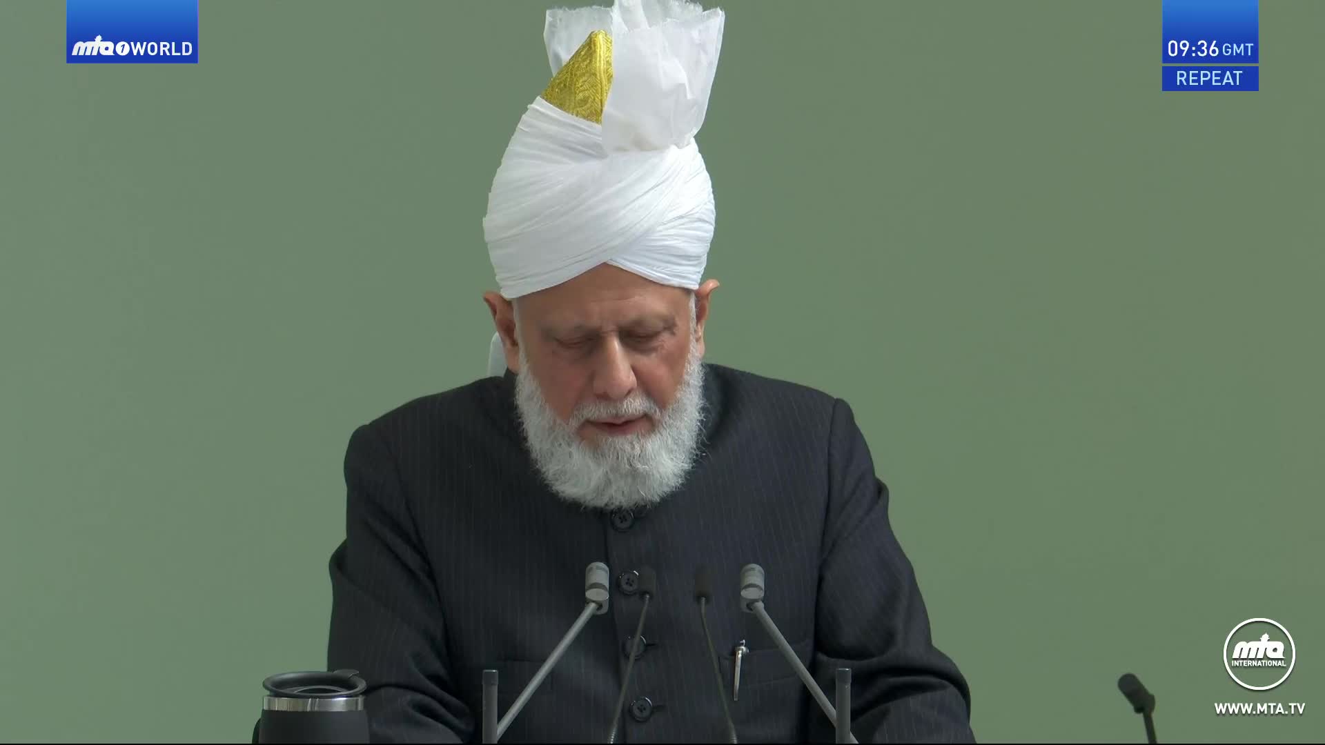 A man with a white turban and beard speaks into two microphones. The screen displays the MTA World logo and the time, 09:36 GMT, with "REPEAT" below it.