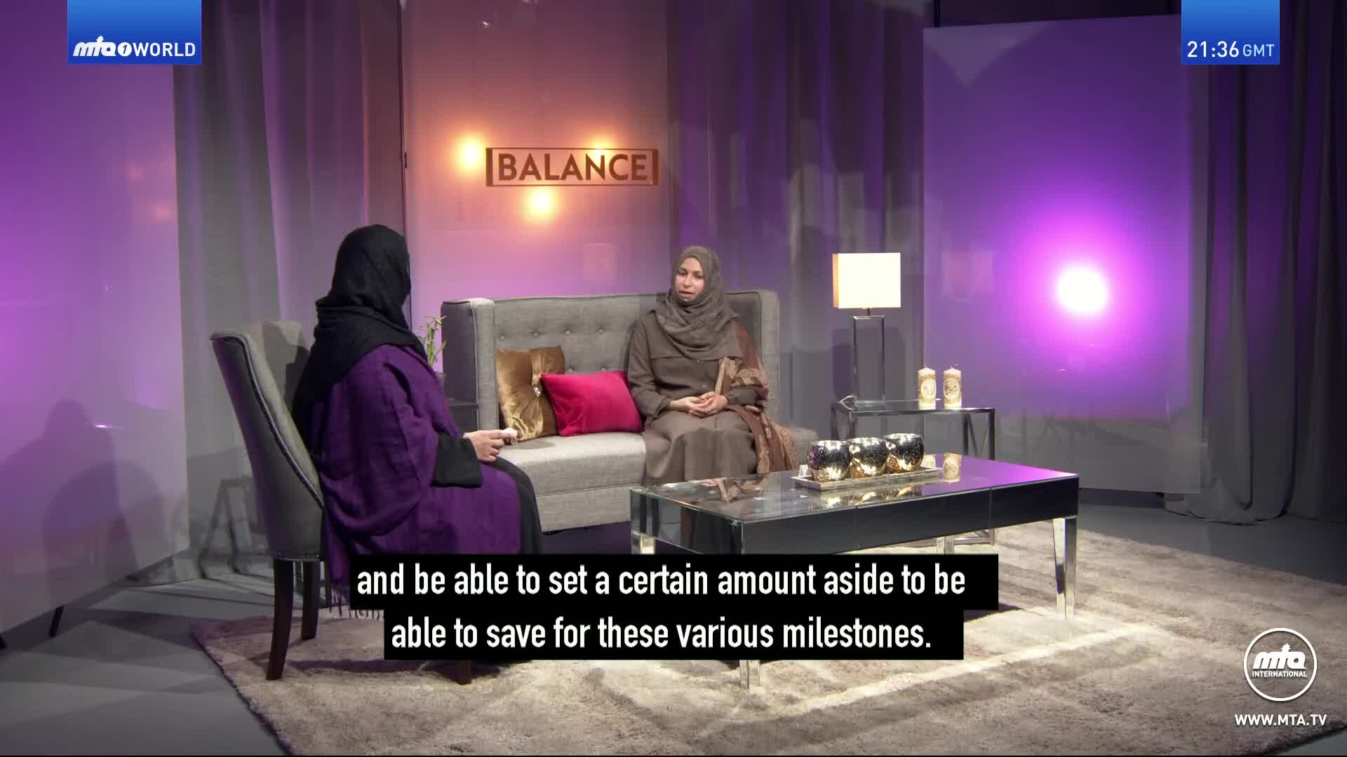 Two women sit on a sofa in a studio, illuminated by soft purple lighting. One woman, facing away from the camera, wears a vibrant purple abaya, while the other, facing the camera, is dressed in a muted brown hijab and dress.