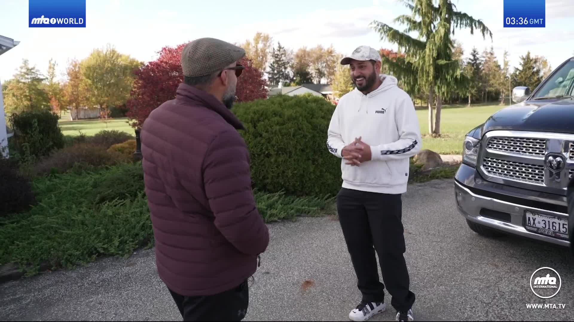Two men stand on a gravel driveway, chatting beside a large black pickup truck. Autumn leaves are scattered across the ground, and the trees in the background are ablaze with red and yellow.