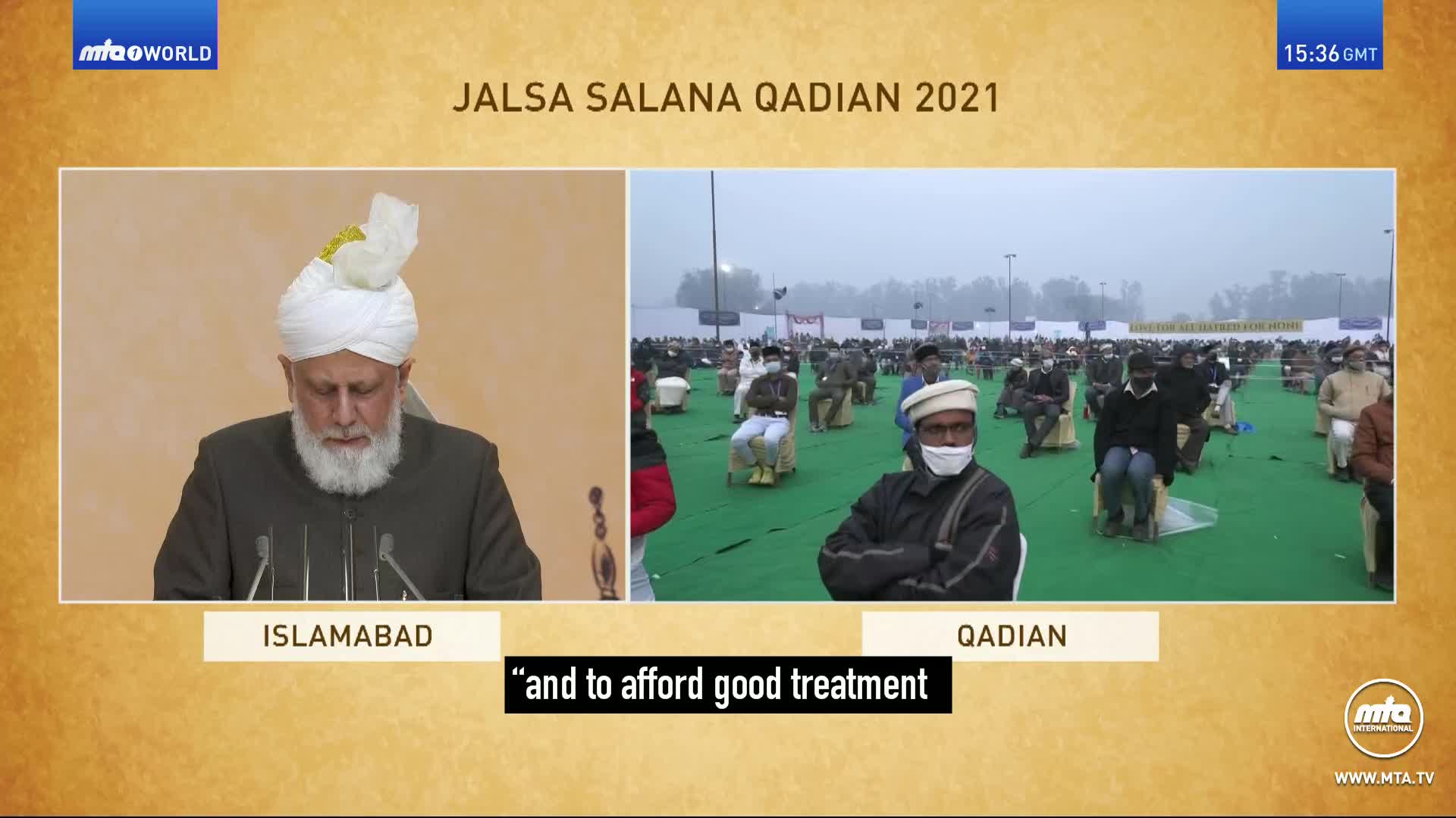 A man in a white turban speaks from a podium, while a crowd sits spaced out on green mats under a hazy sky. The event is the Jalsa Salana Qadian 2021.