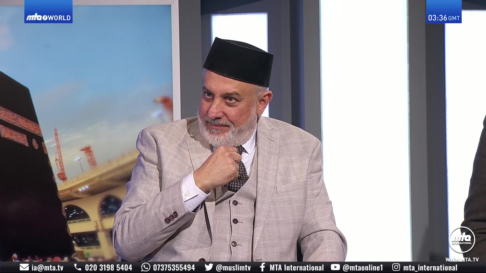 A man in a suit and kufi adjusts his tie. A large screen behind him displays an image of the Kaaba in Mecca.