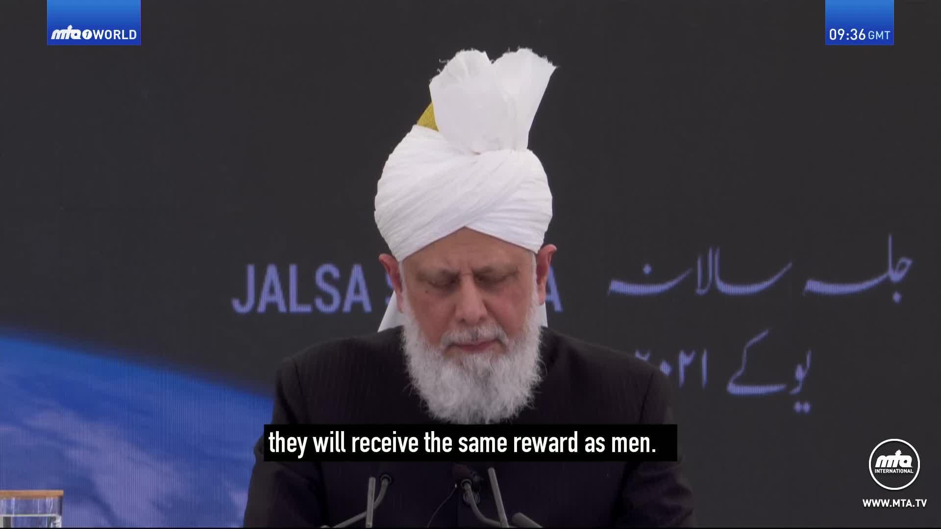 A man in a white turban speaks at a podium. The screen behind him displays the word "JALSA" and Arabic script.