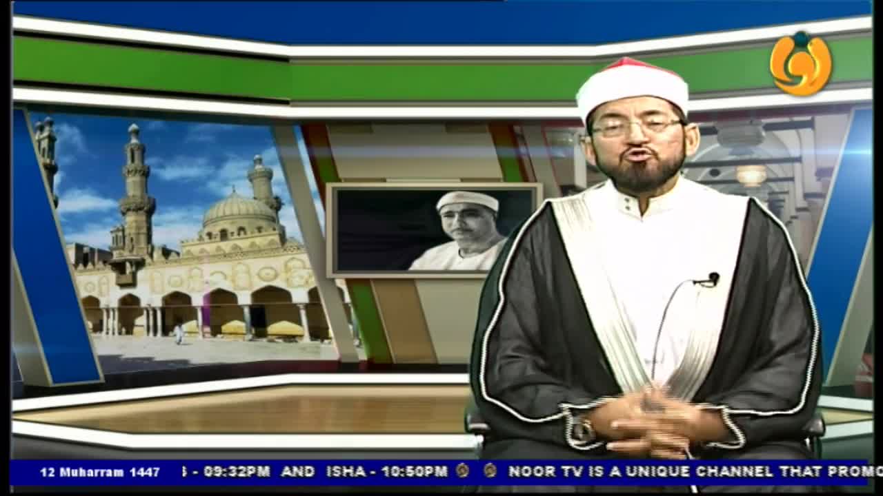 A man in a white turban and dark robes speaks directly to the camera, his hands clasped in his lap. Behind him, a backdrop shows a mosque and a small framed portrait.
