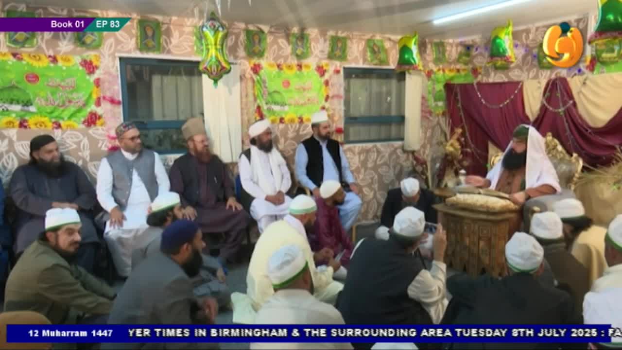 A man with a long beard sits at a lectern, speaking to a group of men gathered on the floor. The scene is being broadcast on Noor TV from Birmingham, United Kingdom.
