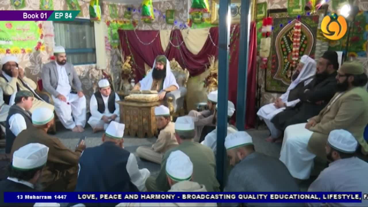 A man with a long beard sits at a small table, speaking to a group of men seated on the floor. The scene is being broadcast by Noor TV, with the United Kingdom in the background.
