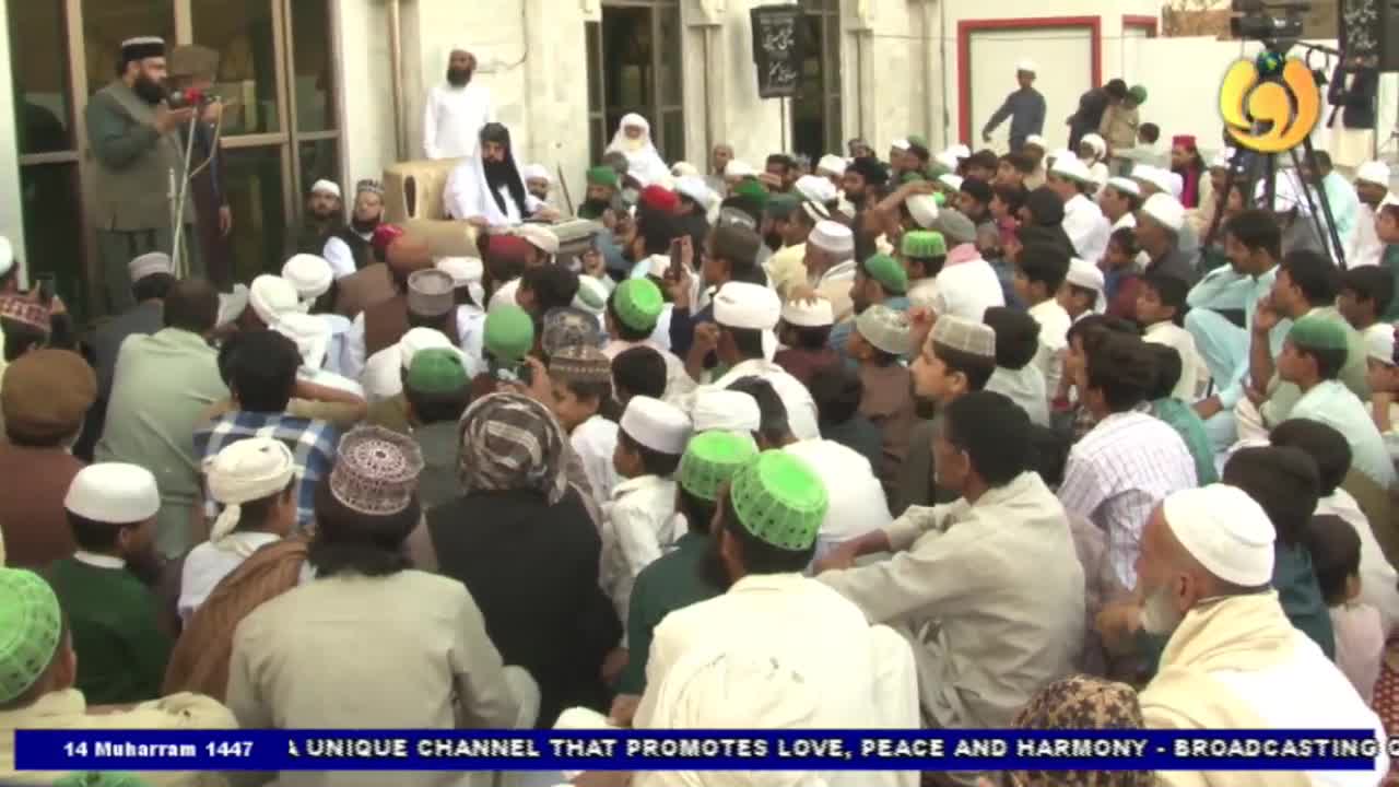 A man with a beard speaks into a microphone, addressing a large crowd seated on the ground. Many in the audience wear white or green caps, and a Noor TV graphic is visible at the bottom of the screen.

