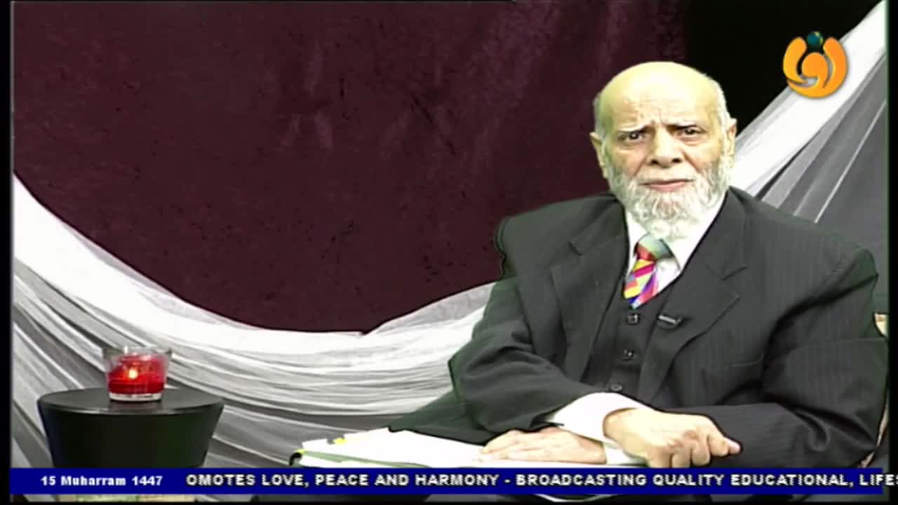 A man in a suit sits at a desk, speaking directly to the camera. A small candle burns on a table to his left, and the Noor TV logo is visible in the corner.

