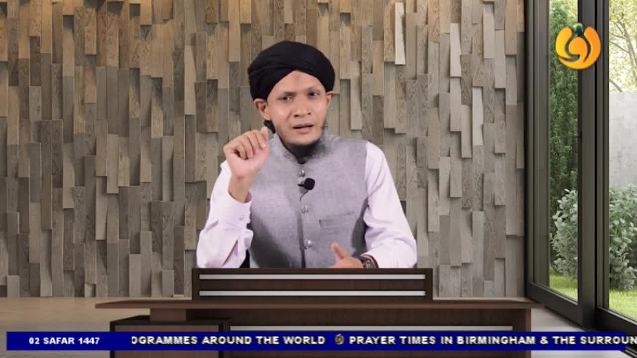 A man in a black turban gestures with his right hand while speaking into a microphone. He's on a set, likely for Noor TV, with a backdrop of wooden panels and a window to the right.
