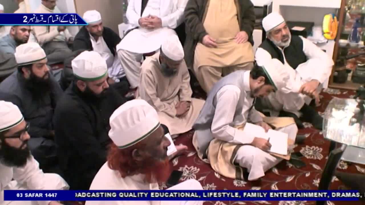 A group of men, most wearing white caps, are gathered on a patterned rug. One man with a long red beard gestures while others listen, and a man in a vest takes notes on a pad of paper.
