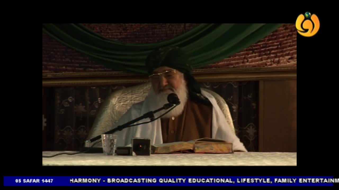 A man with a long white beard speaks into a microphone, his gaze directed forward. On the Noor TV broadcast, a book lies open before him on the table.
