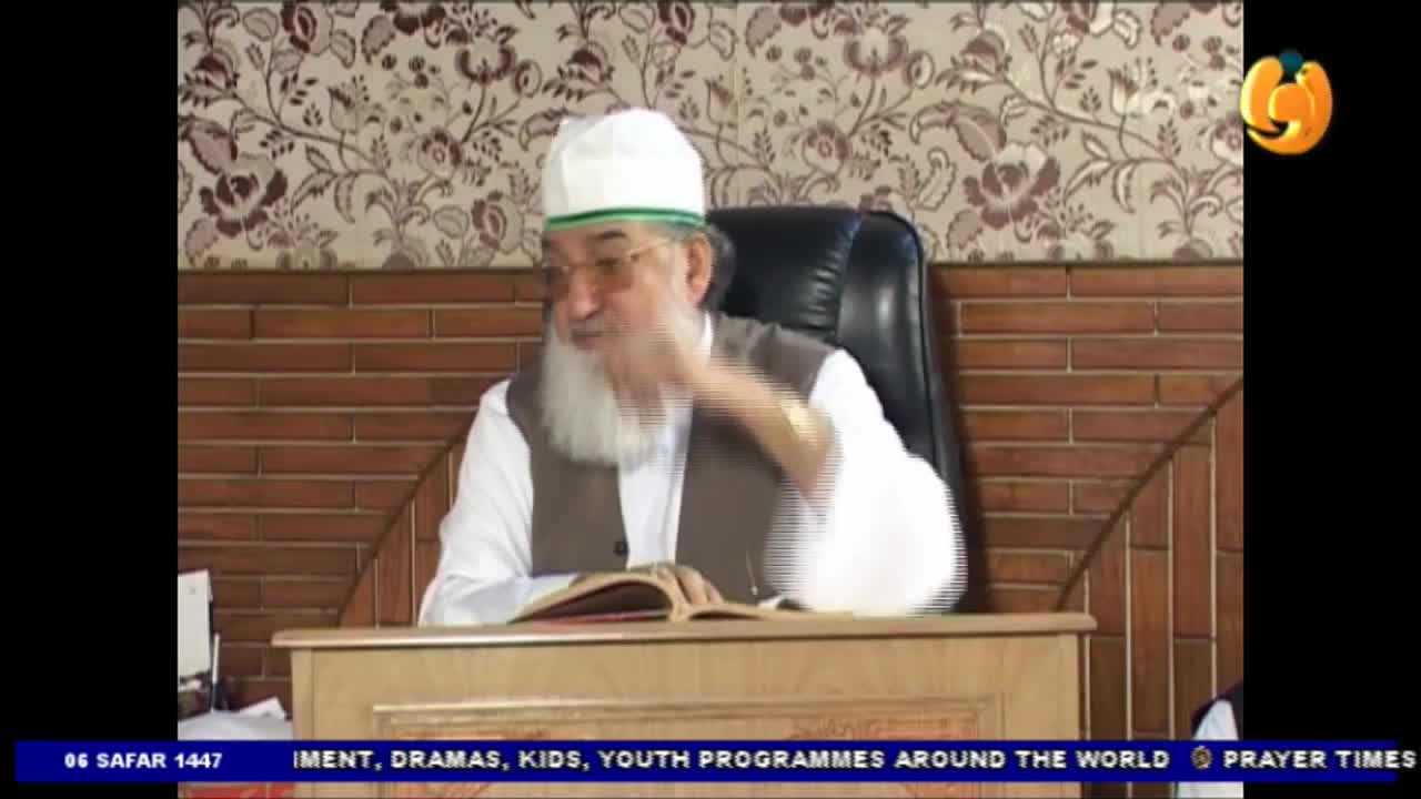 A man with a long white beard gestures with his right hand while speaking from behind a desk. He is wearing a white cap and a brown vest, and a book rests open in front of him.
