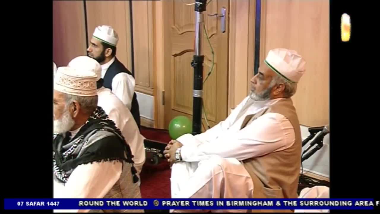 Men in white caps and vests are seated, listening attentively. A Noor TV broadcast is underway, with a banner at the bottom indicating prayer times in Birmingham, United Kingdom.
