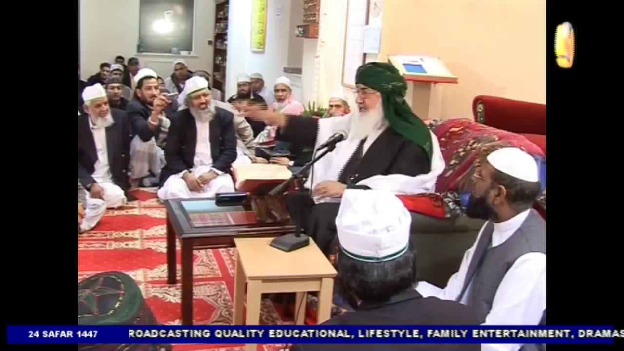 A man in a green turban gestures with his right hand as he speaks into a microphone. Seated on the floor, the audience listens intently, some wearing white caps.
