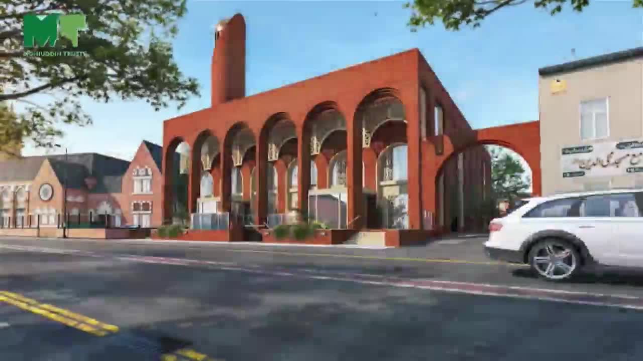 A modern mosque with a tall, curved minaret stands on the corner, its red brick facade contrasting with the blue sky. A white car drives past, and the street is lined with other buildings.
