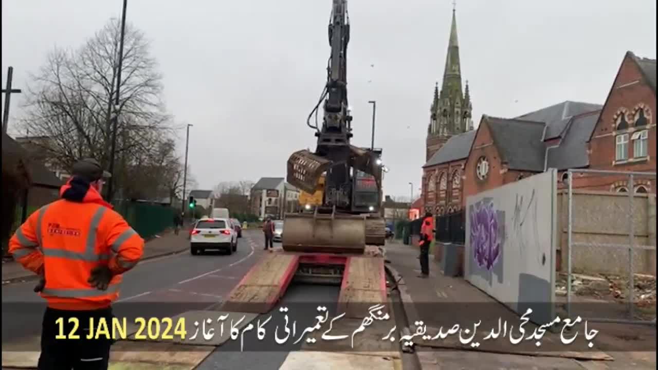 A large excavator is being unloaded from a flatbed trailer on a street in the United Kingdom. The date, 12 January 2024, is displayed, along with Urdu text, possibly related to a construction project.
