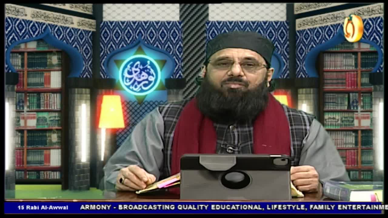 A man with a long, dark beard sits behind a desk, facing the camera. He's wearing a black cap and a red scarf, with a tablet in front of him, likely on a Noor TV broadcast from the United Kingdom.
