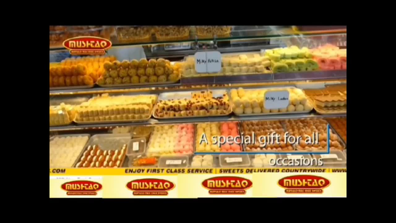 A brightly lit display case is filled with various sweets, each type neatly arranged in rows. The Noor TV advertisement highlights the "Mushtaq" brand, promising sweets delivered nationwide.
