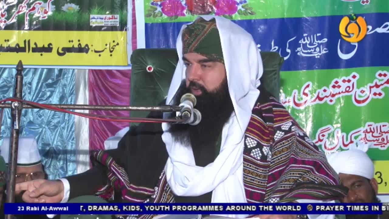 A man with a long black beard speaks into a microphone, his hands gesturing as he addresses the audience. He wears a turban and a patterned shawl, with the Noor TV logo visible in the corner.
