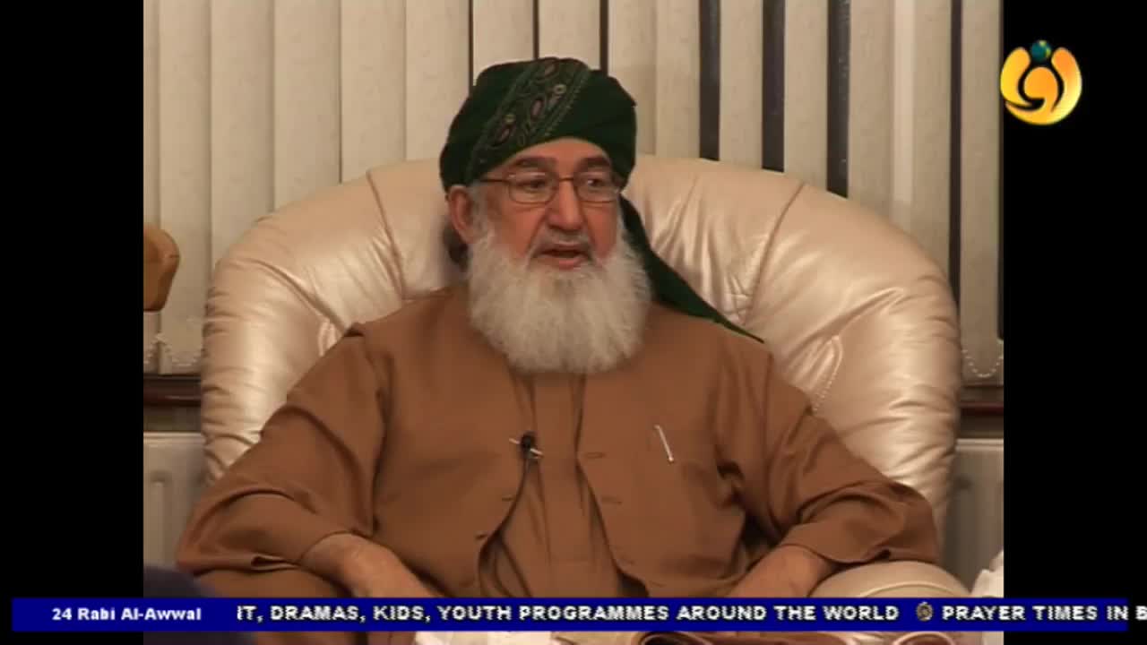 A man wearing a green turban and glasses sits in a large, cream-colored armchair. He's speaking, and a microphone is clipped to his collar.
