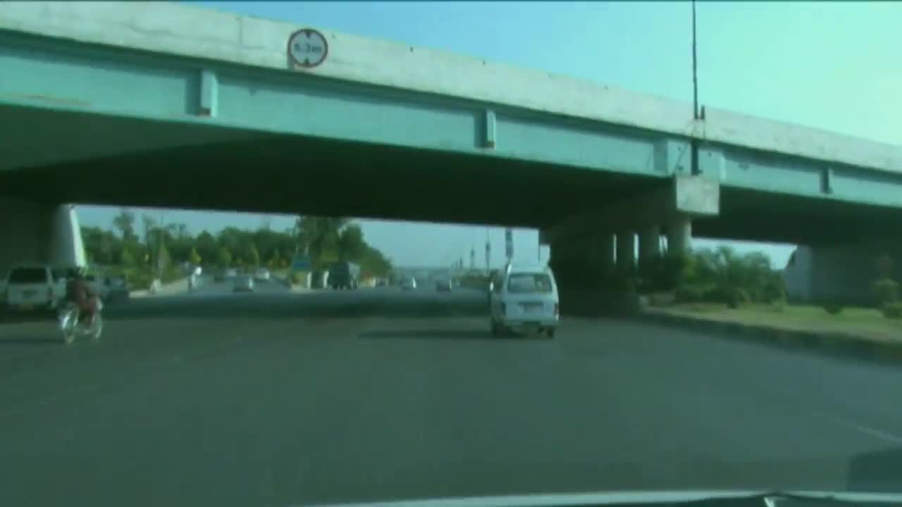Beneath a concrete overpass, a white van drives down the road, followed by a motorcycle. The sky is clear, and the sun is shining.

