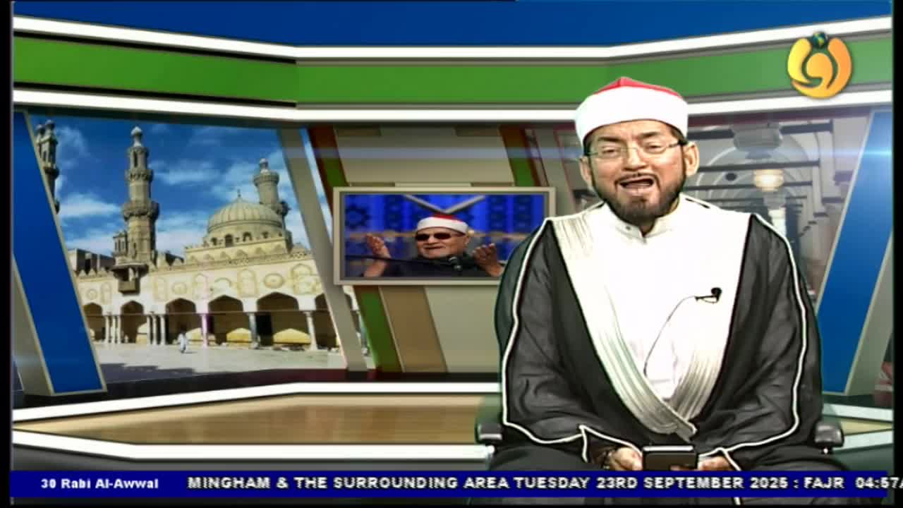 A man in a white turban and traditional robes smiles, speaking into a microphone on a television set. Behind him, a smaller screen shows another man, and a backdrop depicts a mosque.
