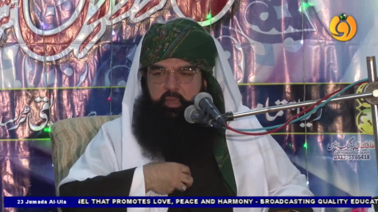 A man with a long black beard and a green turban sits at a lectern, speaking into two microphones. Behind him, a backdrop displays ornate patterns and text, likely for a Noor TV broadcast.
A man with a long black beard and a green turban sits at a lectern, speaking into two microphones. Behind him, a backdrop displays ornate patterns and text, likely for a Noor TV broadcast.