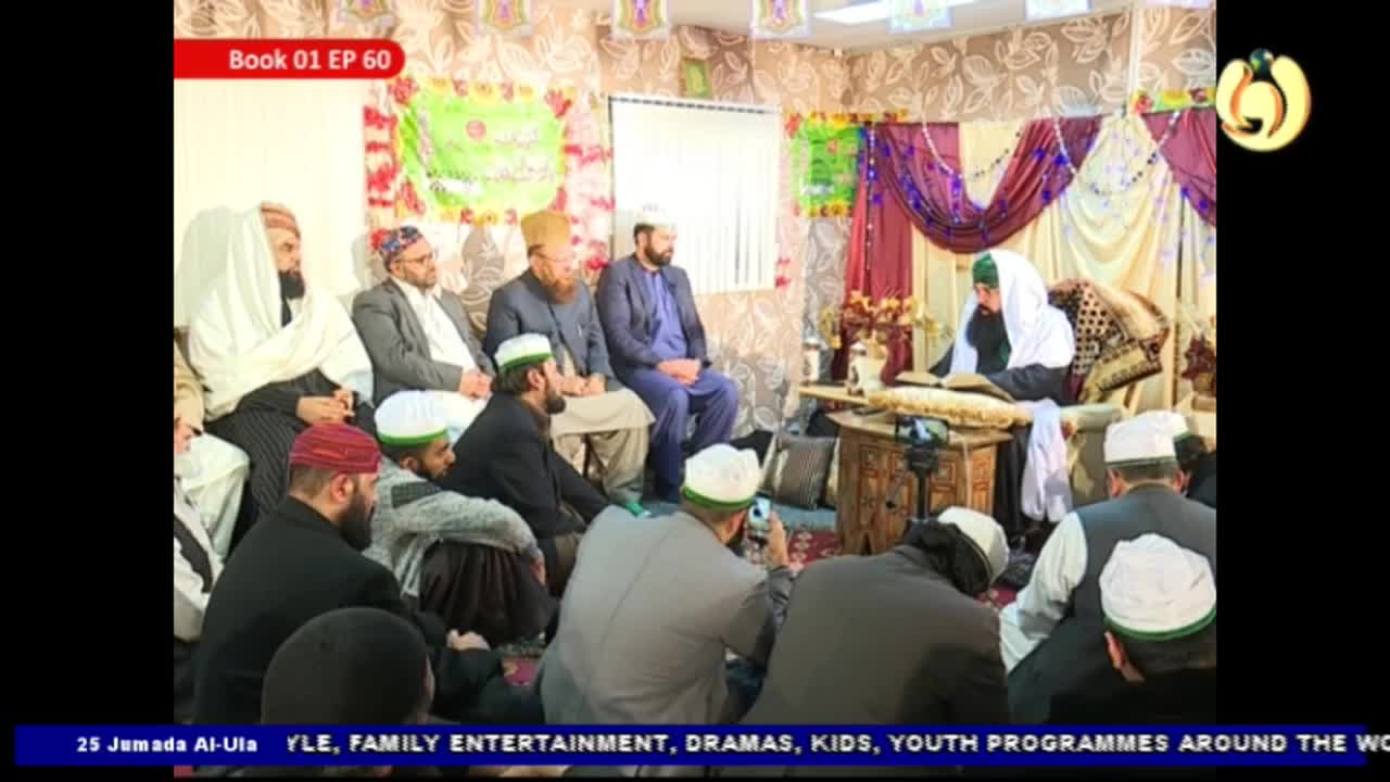 A robed figure reads from a book at a small table, facing a seated audience. The scene appears to be broadcast on Noor TV, with the United Kingdom likely being the location.
A robed figure reads from a book at a small table, facing a seated audience. The scene appears to be broadcast on Noor TV, with the United Kingdom likely being the location.