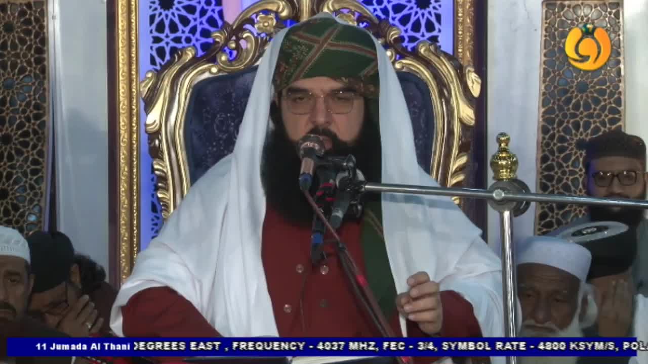A man in a turban and white shawl speaks into a microphone, his words likely broadcast by Noor TV.  He's seated in an ornate chair, with others gathered nearby.

