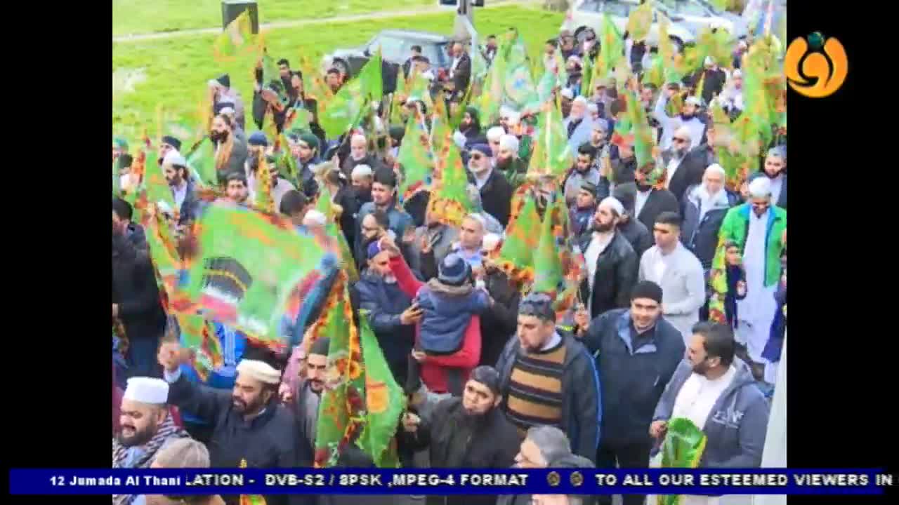A crowd of men, many with white caps and beards, are walking forward, waving green flags. A small child is held aloft in the middle of the procession.
