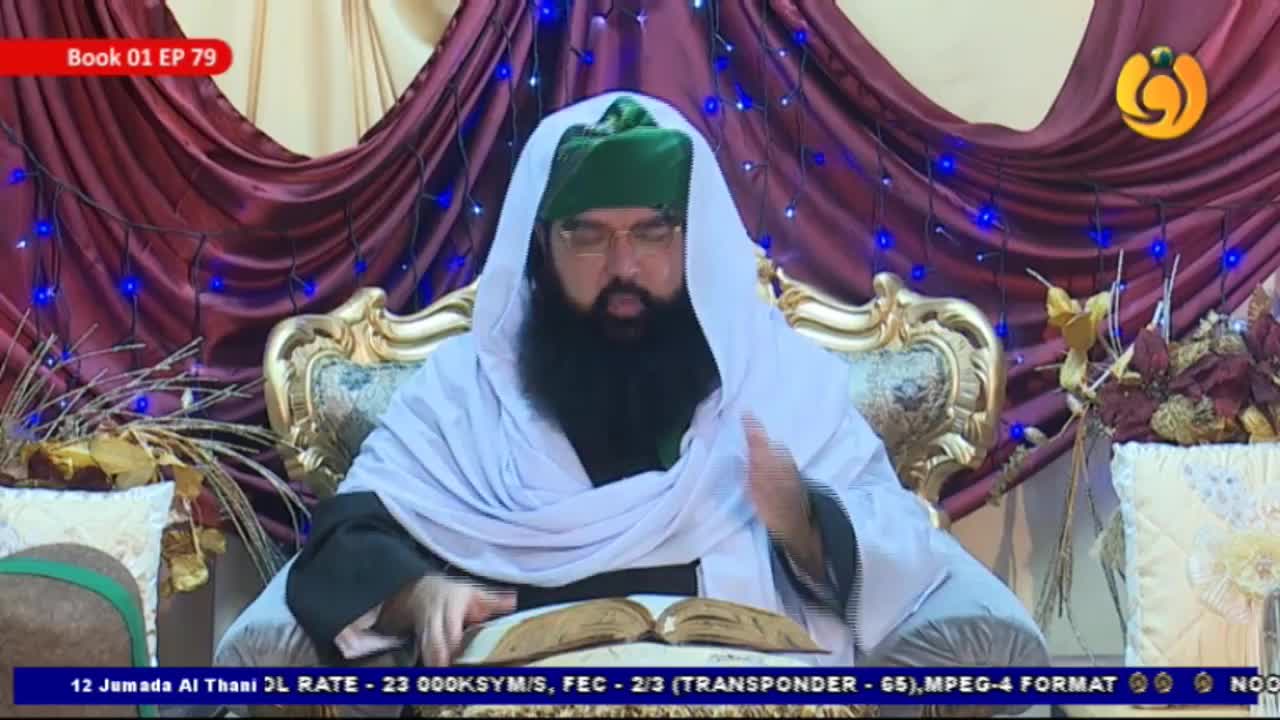 A man in a green turban and white shawl sits in an ornate chair, gesturing with his right hand as he speaks. He's reading from a large, open book, likely for a broadcast on Noor TV from the United Kingdom.
