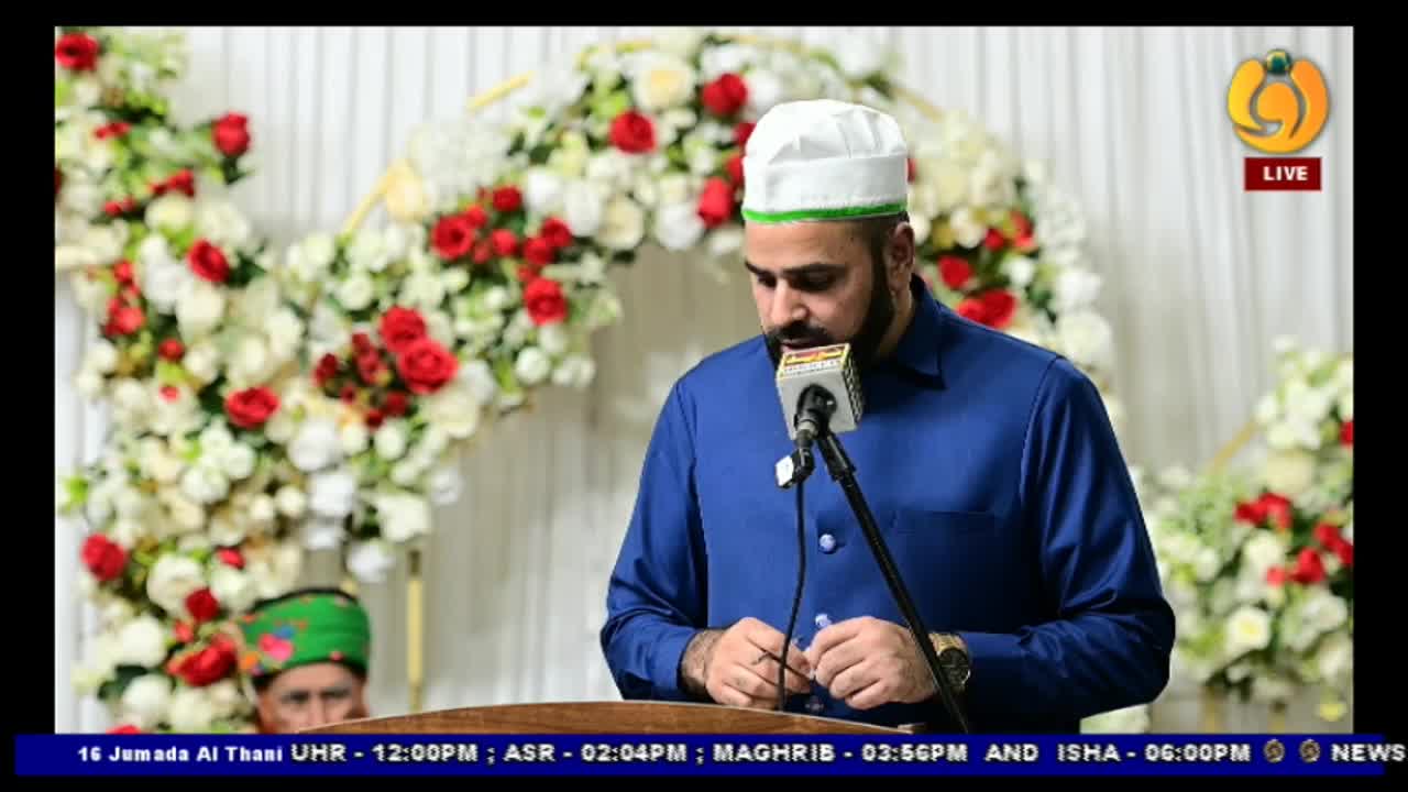 A man in a blue suit and white cap speaks into a microphone, likely on Noor TV, with a floral arch as a backdrop. Below the screen, a ticker displays the time and prayer schedules.
