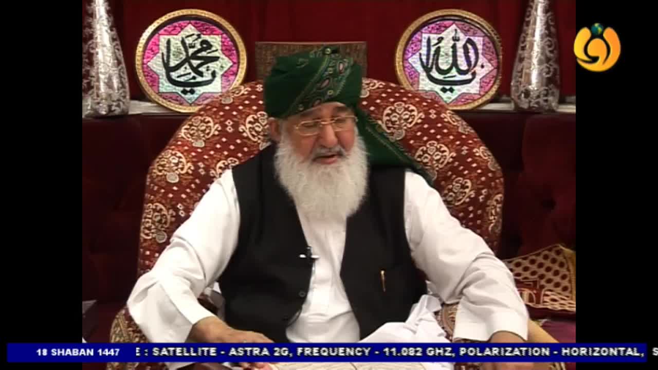 An older man with a long white beard, wearing a green turban and a dark vest over a white shirt, sits on a plush seat. He is speaking, his head tilted slightly as if addressing an audience, with ornate Islamic calligraphy displayed on the wall behind him.