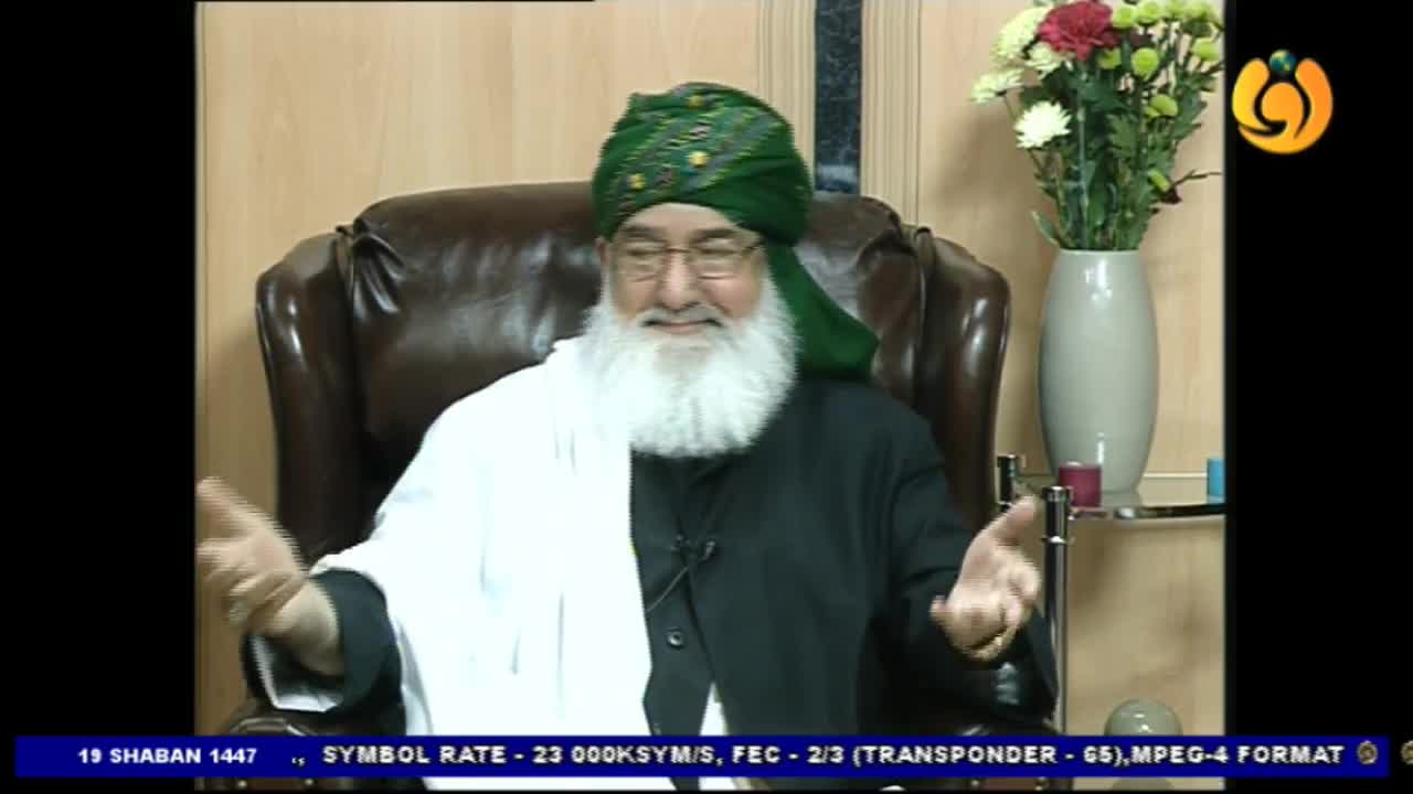 A man in a green turban and white shawl gestures with his hands while speaking. He sits in a dark leather chair, a vase of flowers visible behind him.
