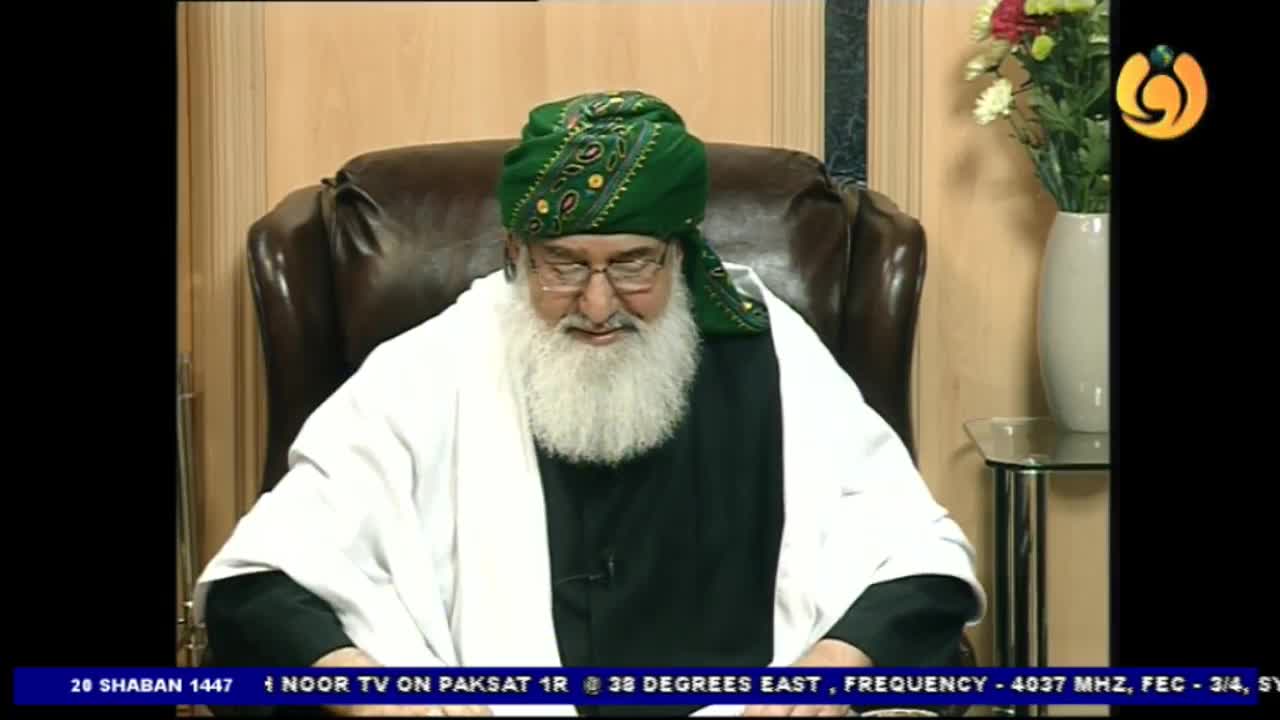 A man with a long white beard and a green turban sits in a leather chair, looking down. He wears a white robe over a black garment, broadcasting on Noor TV.