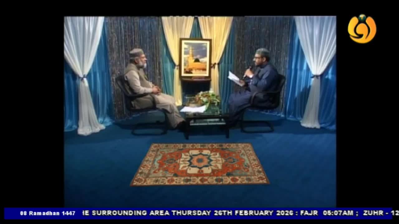 Two men sit facing each other on a blue carpeted set, one holding a microphone and a paper. Behind them, draped blue and gold curtains frame a picture of a mosque. Two men sit facing each other on a blue carpeted set, one holding a microphone and a paper. Behind them, draped blue and gold curtains frame a picture of a mosque.
