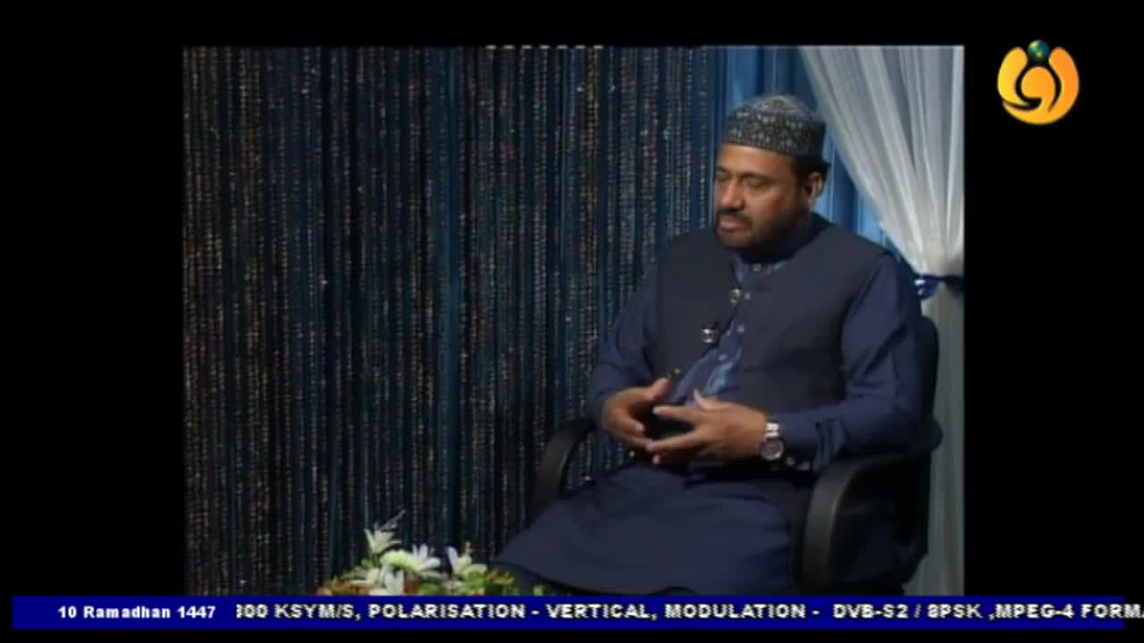 A man in a dark blue outfit and a patterned cap sits in a chair, speaking. He gestures with his hands as he talks, his watch glinting under the studio lights. A man in a dark blue outfit and a patterned cap sits in a chair, speaking. He gestures with his hands as he talks, his watch glinting under the studio lights.
