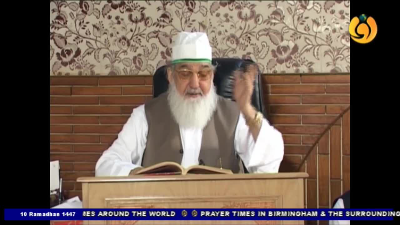 An older man with a long white beard, wearing a white cap and vest over a white robe, gestures with his right hand as he speaks. He is seated behind a wooden desk with an open book before him, likely during a broadcast for Noor TV. An older man with a long white beard, wearing a white cap and vest over a white robe, gestures with his right hand as he speaks. He is seated behind a wooden desk with an open book before him, likely during a broadcast for Noor TV.